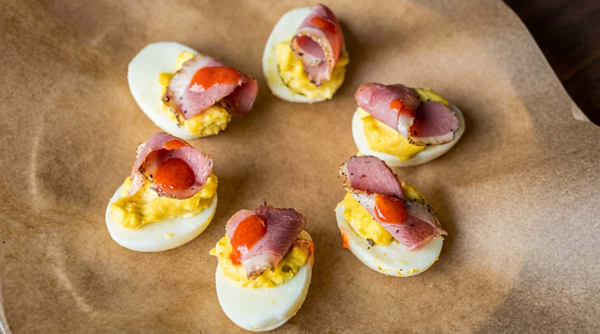 Six deviled eggs with mustard-colored filling, topped with folded ham and red sauce, on brown parchment paper
