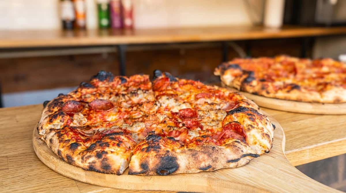 Pepperoni pizza with charred crust, melted cheese, and tomato sauce, on a wooden board with a neon "Let's Party" sign in the background.
