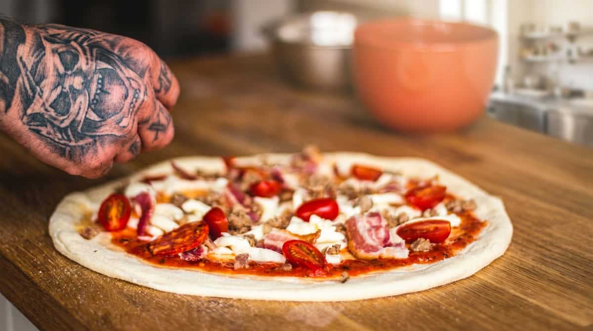 Uncooked pizza with cherry tomatoes, mozzarella, bacon, sausage, and pepperoni, prepared by a tattooed hand.