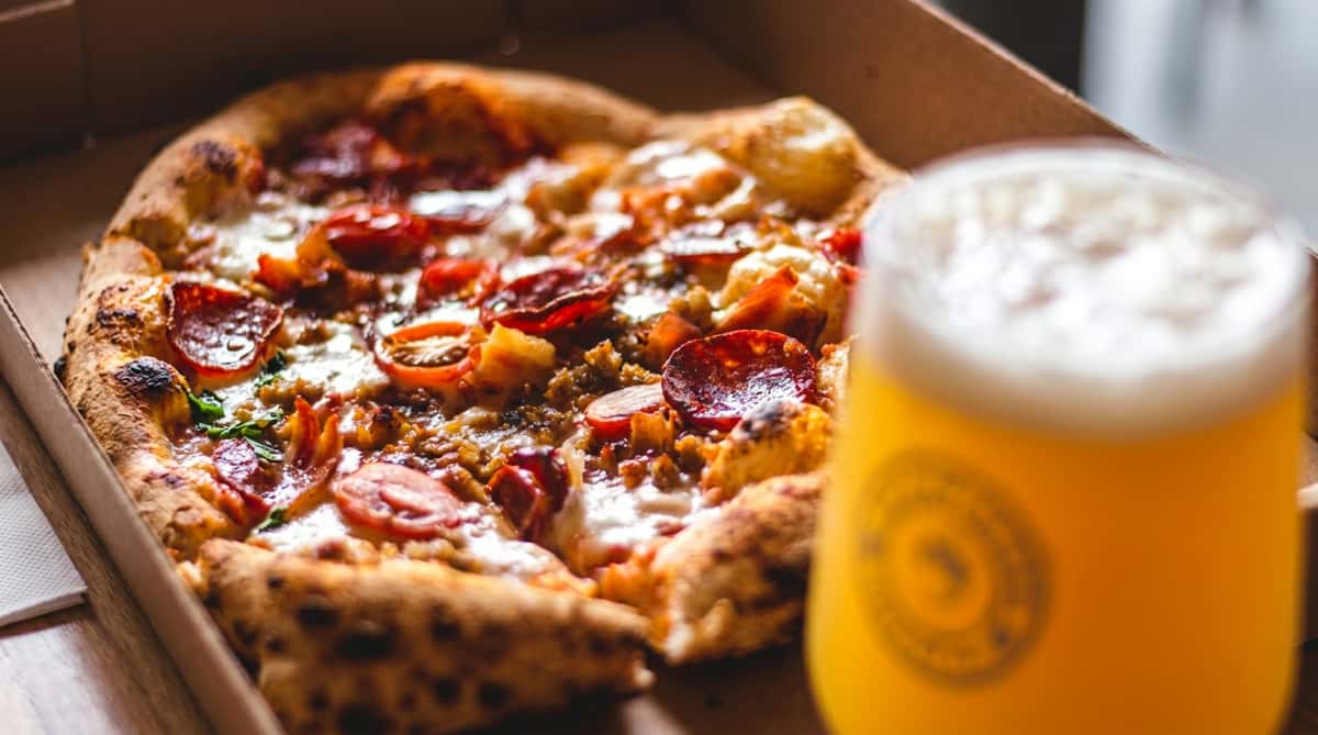 Pepperoni pizza with sausage and melted cheese in a box, with a glass of beer in the foreground.