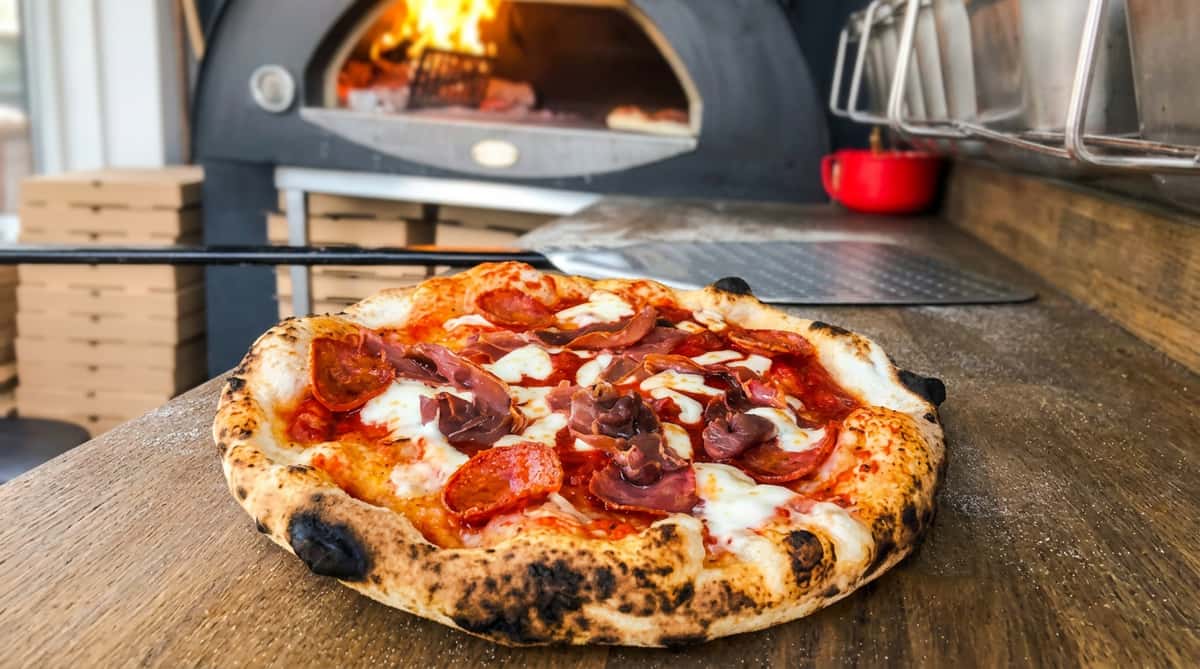 Neapolitan pizza with pepperoni, prosciutto, and mozzarella on a wooden peel, in front of a wood-fired oven.