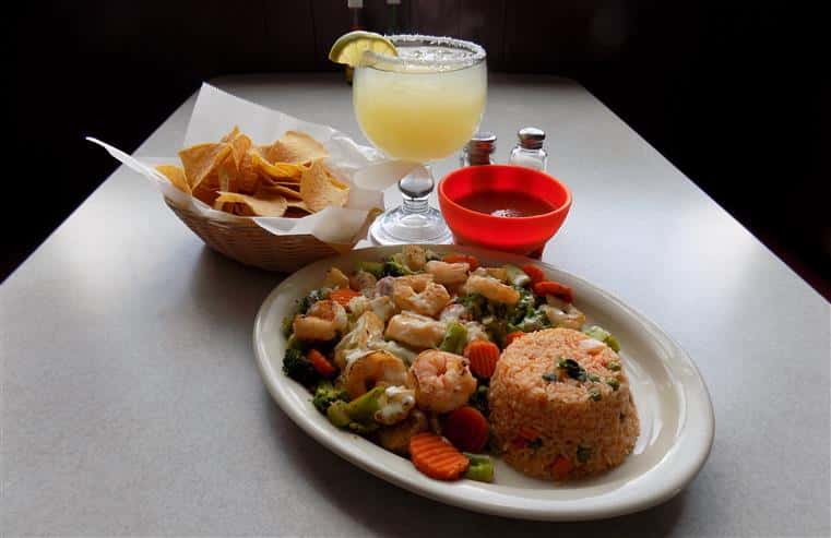 plate with cooked shrimp, a variety of vegetables and rice. On the side is a cup of salsa and a basket of chips, and a margarita with a salt rim and lime slice
