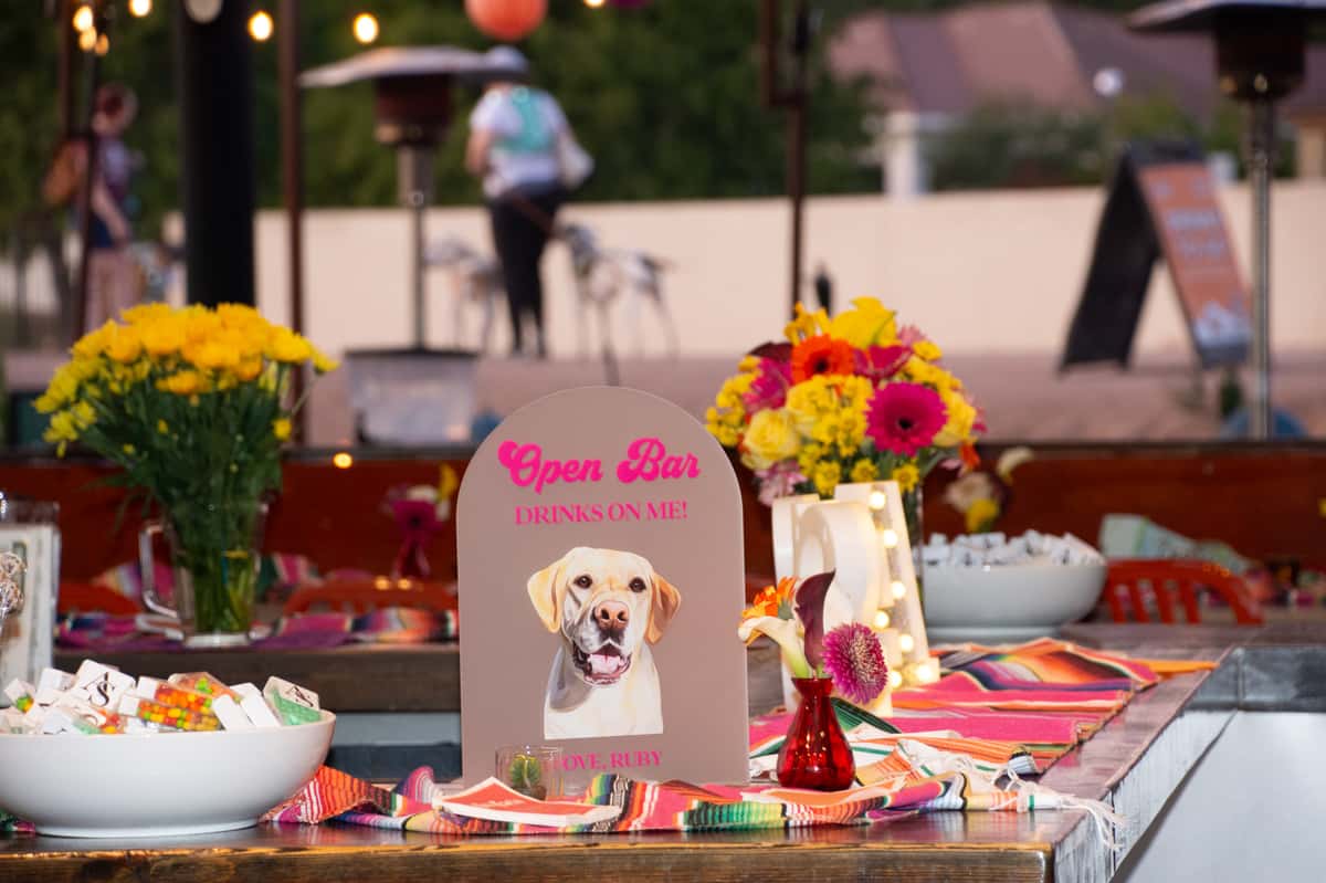 Bar with decor of flowers and mexican blankets and a photo of a dog that says drinks on me