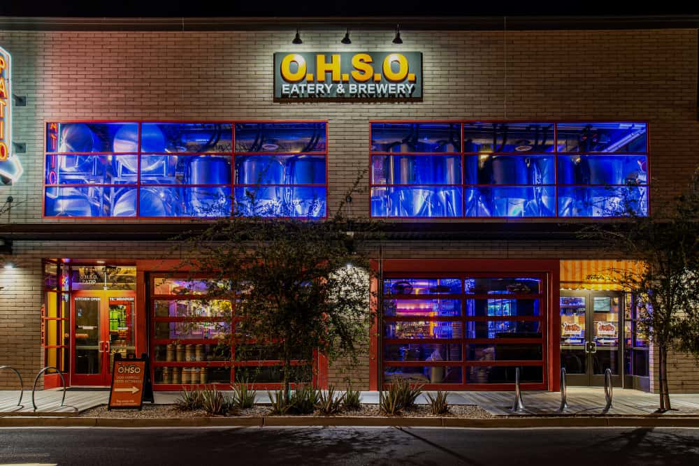 Exterior of O.H.S.O. Gilbert in Downtown Gilbert at night, featuring the two-story brewery and restaurant with brewing tanks visible through the windows.