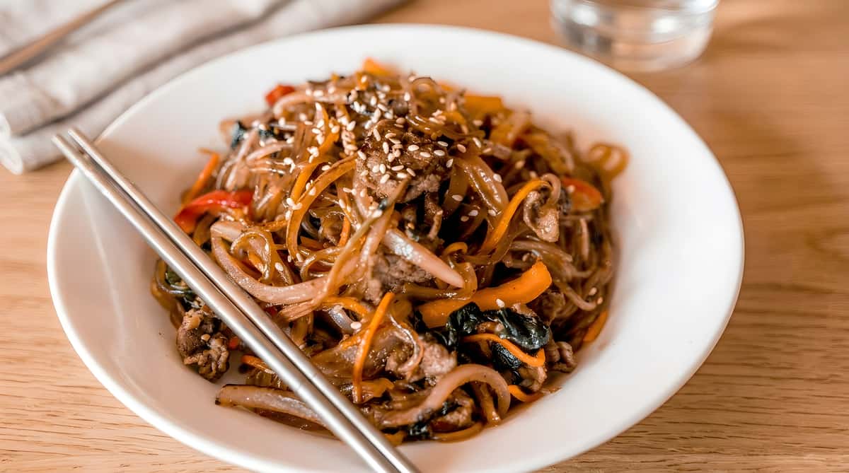Japchae with glass noodles, sliced beef, carrots, onions, and sesame seeds; chopsticks rest on the dish.