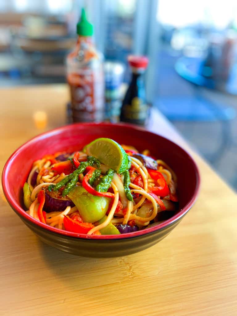 Colorful noodle bowl with bok choy, red peppers, and lime, topped with green sauce, set on a wooden table in a casual eatery.