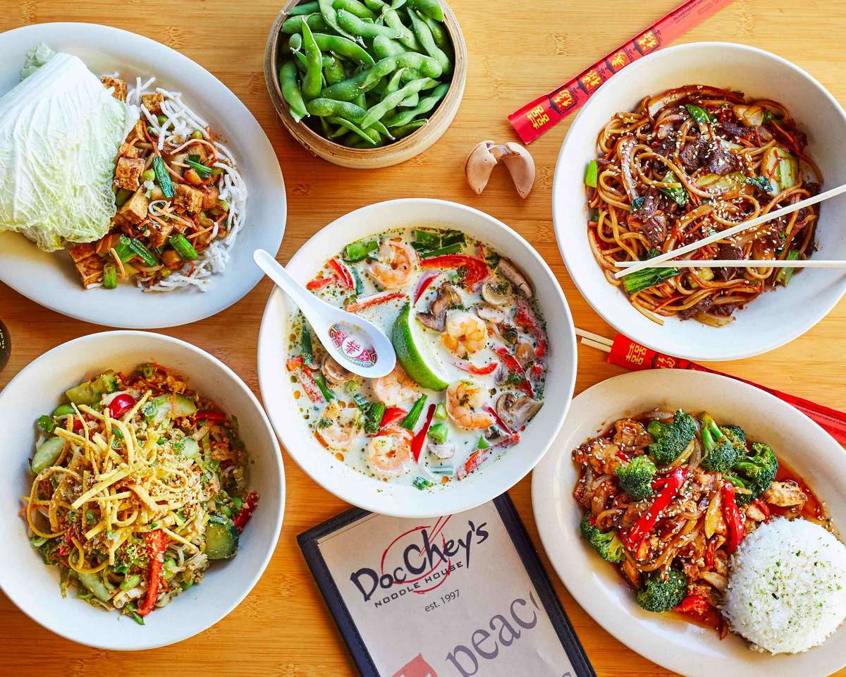 Colorful spread of Asian dishes from Doc Chey's, featuring noodles, stir-fry, curry soup, lettuce wraps, edamame, and rice.