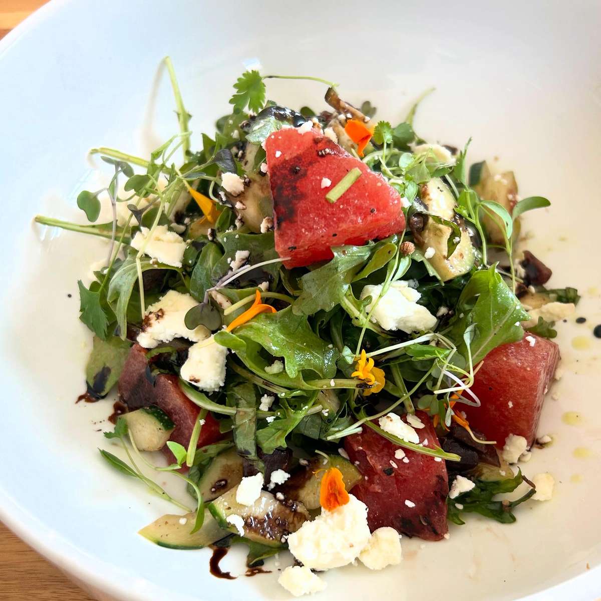 A refreshing salad with watermelon, mixed greens, cucumber, goat cheese, microgreens, and edible flowers, drizzled with balsamic vinaigrette.