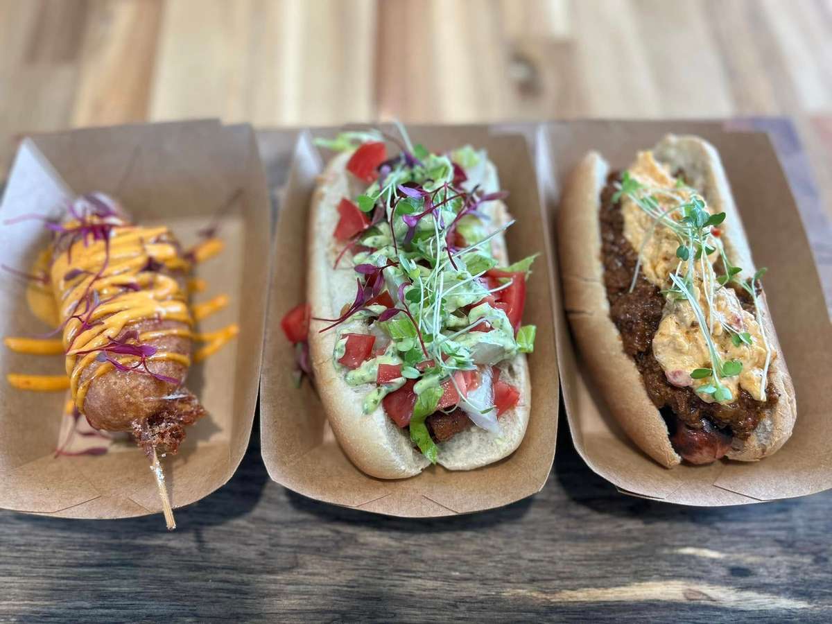 Trio of gourmet hot dogs: a corn dog with spicy aioli, a loaded veggie dog, and a chili cheese dog with microgreens.