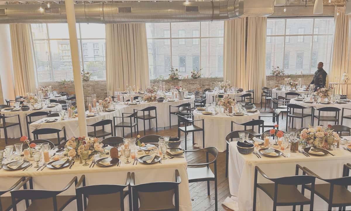 Elegant event dining setup with round tables, white linens, floral centerpieces, and fine china in a modern, industrial venue, with natural light streaming through large windows.