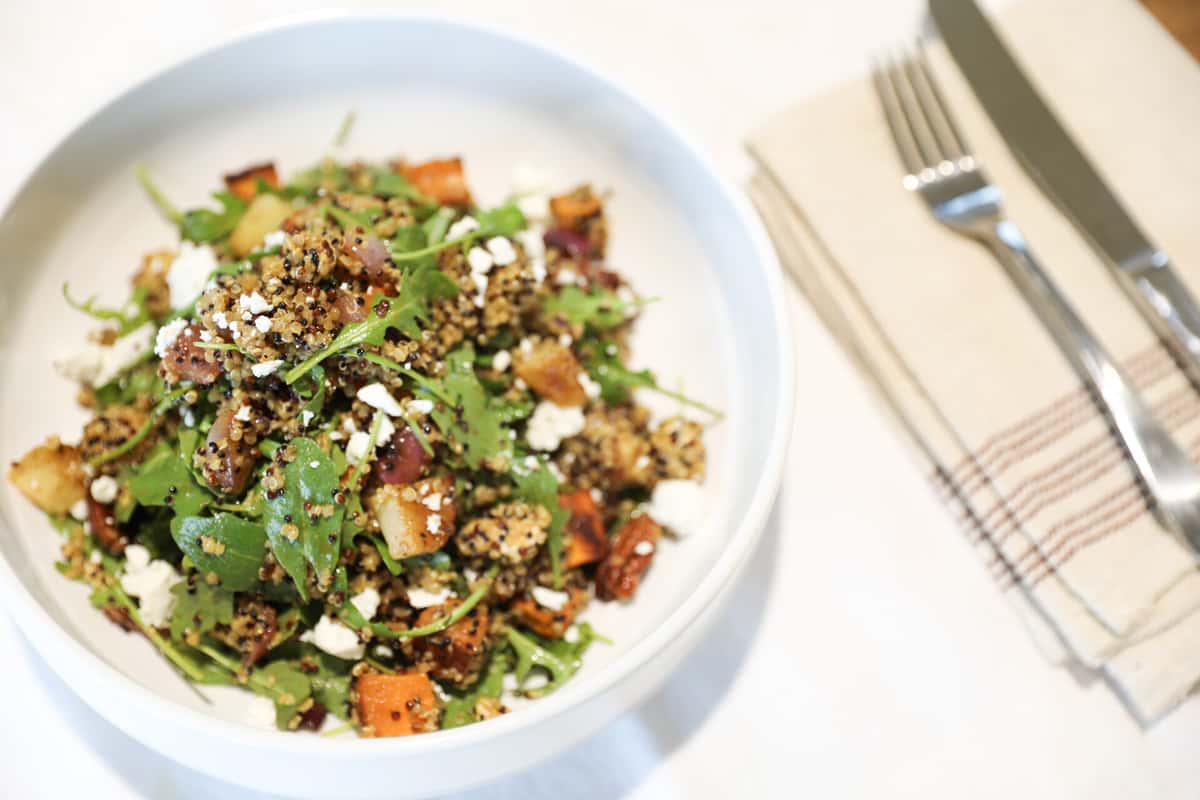 A fresh salad featuring quinoa, roasted sweet potatoes, arugula, and crumbled feta cheese, served in a white bowl.