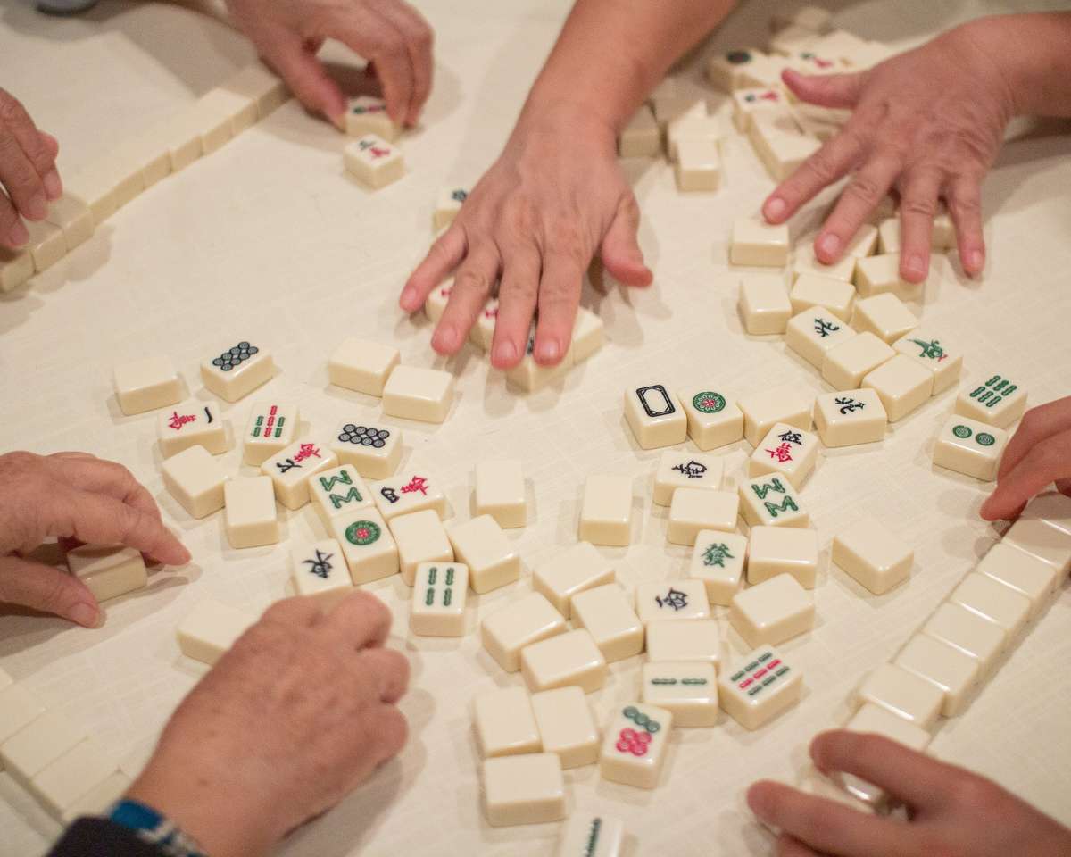 A close-up of several hands playing a game of Mahjong, with tiles arranged on a table in a casual, competitive setting.
