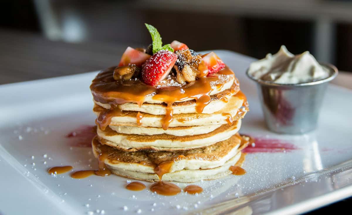 Stack of fluffy pancakes drizzled with rich caramel sauce, topped with fresh strawberries, caramelized bananas, a mint sprig, and a dusting of powdered sugar, served with a side of whipped cream on a white rectangular plate.