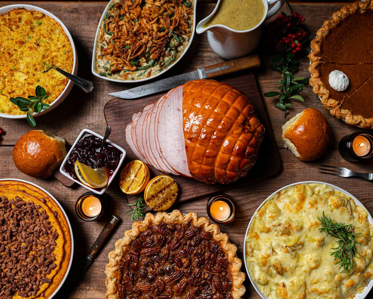 Overhead view of a festive meal centered on glazed sliced ham, surrounded by casseroles, gravy, rolls, cranberry sauce, pumpkin and pecan pies, and candles.
