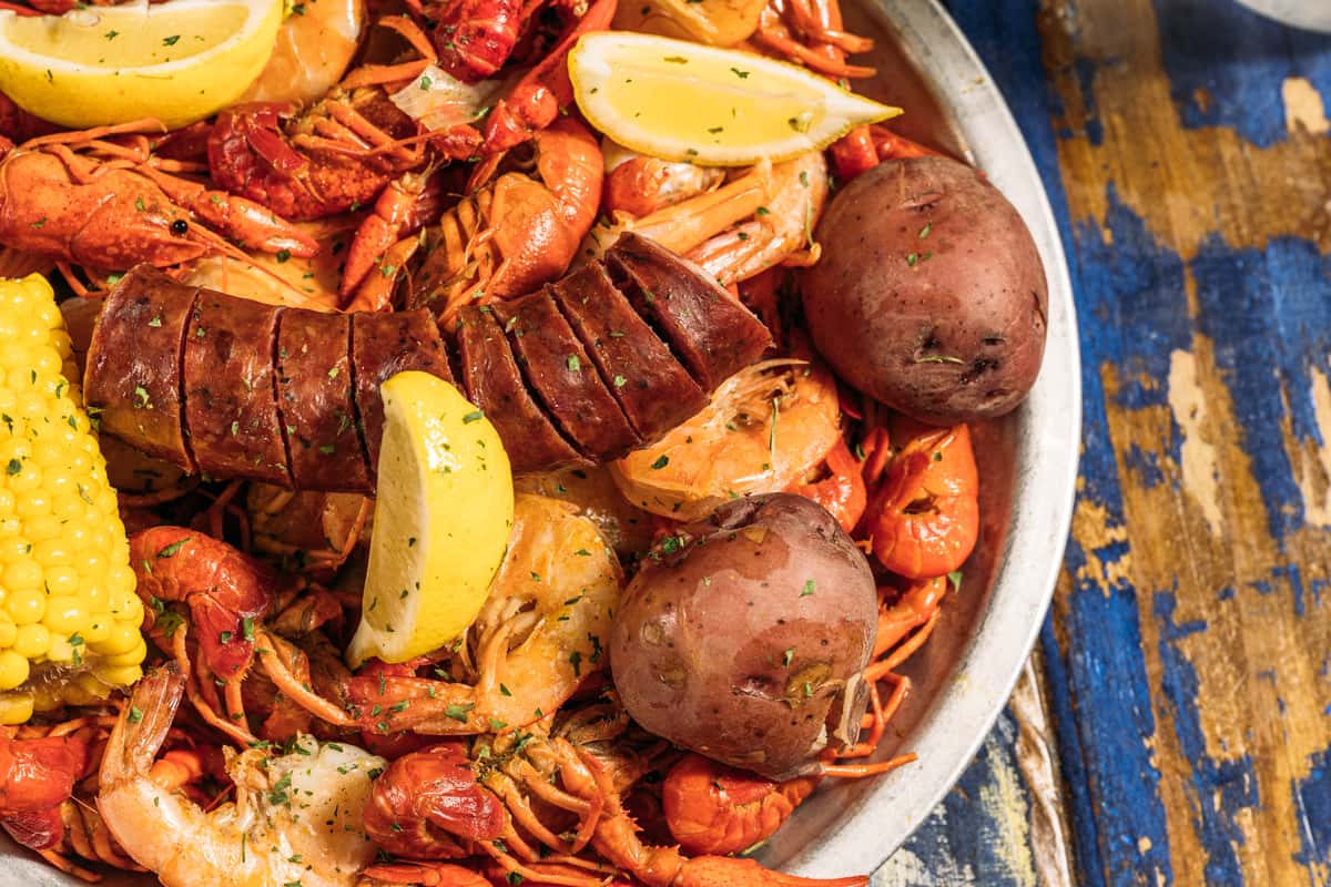 Close-up of a Cajun seafood boil with seasoned crawfish, sliced andouille sausage, red potatoes, corn on the cob, shrimp, and lemon wedges served in a metal bowl on a rustic wooden table.