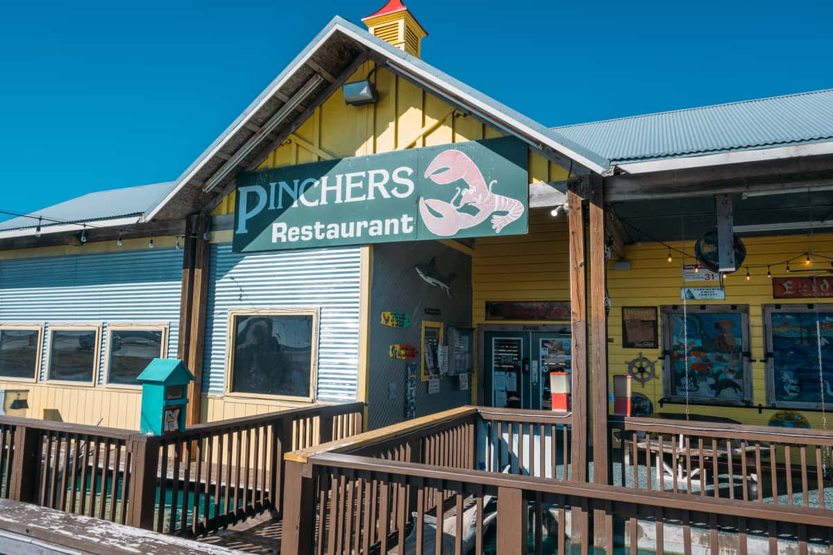 Close-up of the Pinchers Restaurant entrance with wooden deck, lobster logo sign, yellow siding, and marina-style design