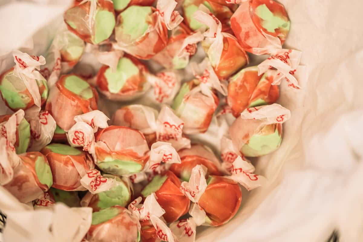Saltwater taffy candies in assorted pastel orange and mint green flavors, individually wrapped in wax paper, displayed in a bulk bin at a nostalgic candy shop or dessert counter.