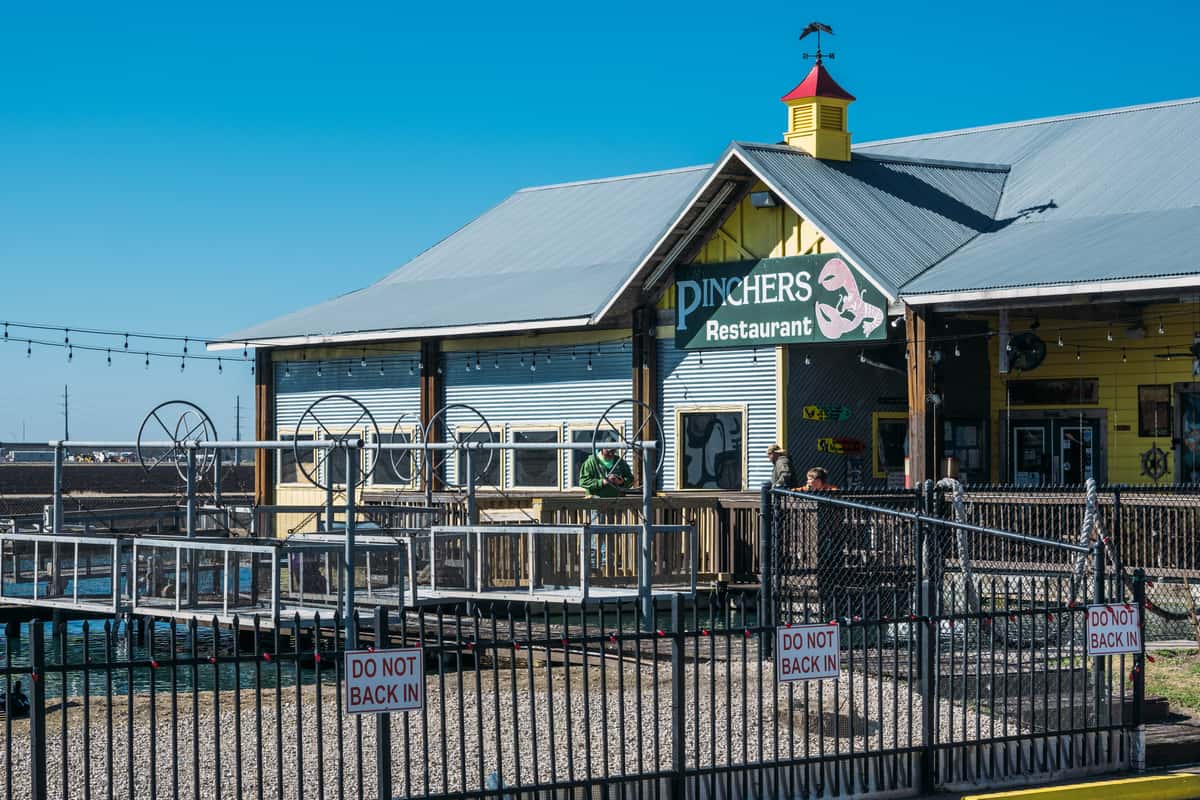 Exterior view of Pinchers Restaurant, a waterfront seafood restaurant with dockside seating, metal siding, and marina views under a clear blue sky, highlighting casual coastal dining.