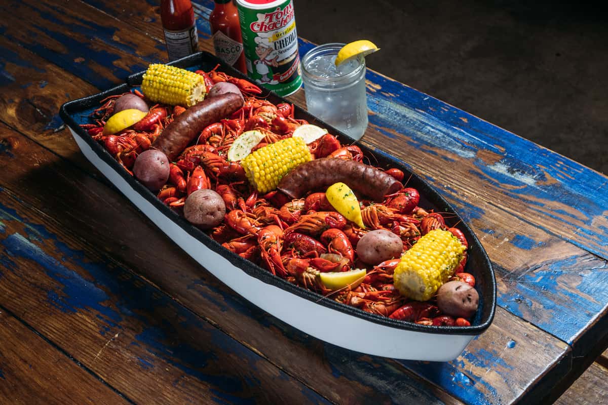 Cajun seafood boil with crawfish, smoked sausage, corn on the cob, red potatoes, and lemon wedges served in a boat-shaped tray, paired with hot sauce and a lemon-garnished cocktail on a rustic wooden table, showcasing Southern Louisiana cuisine and casual restaurant ambiance.