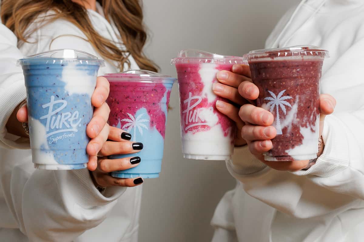 Close-up of hands holding three vibrant Pure Superfood smoothies in blue, magenta, and dark acai hues, swirled with creamy white yogurt.
