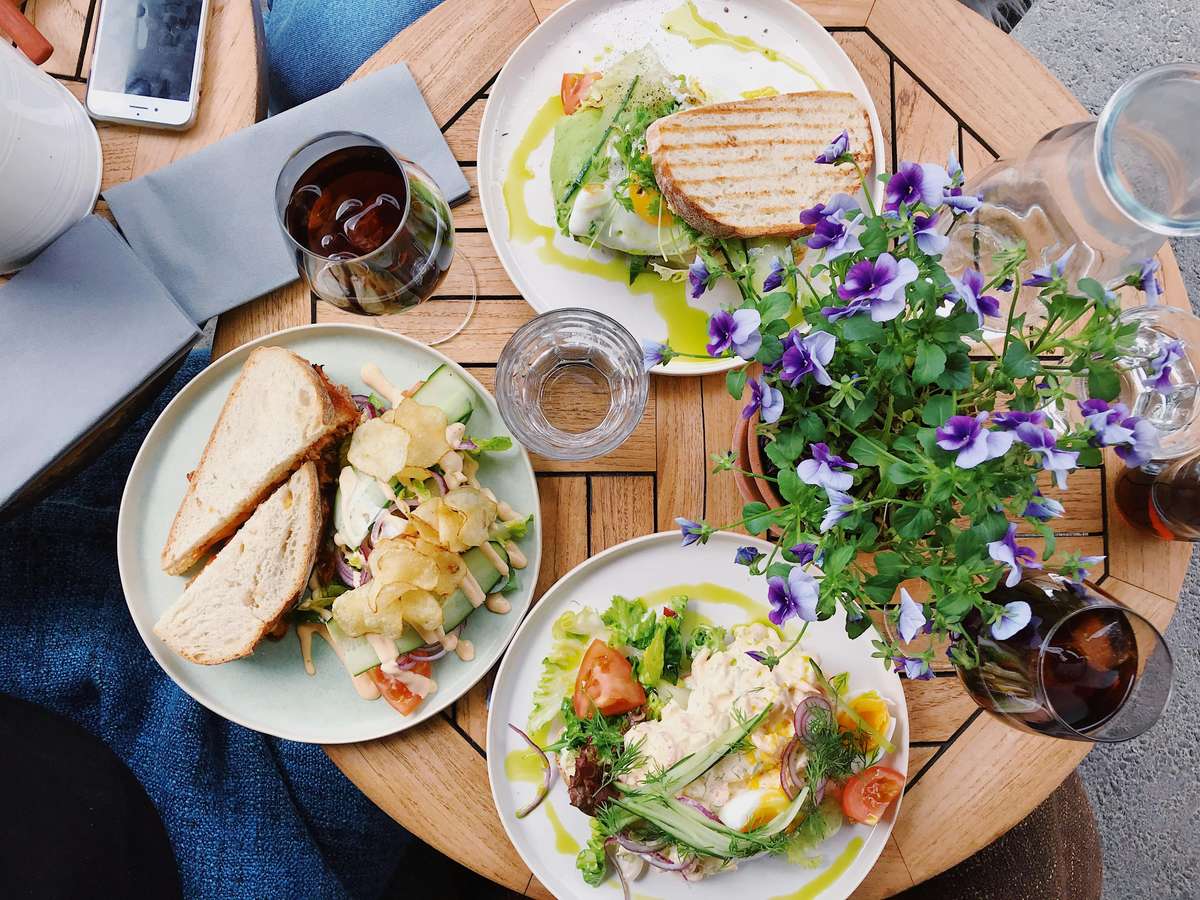 Outdoor cafe table with sandwiches, fresh salads, drinks, and a vase of purple flowers on a wooden table.