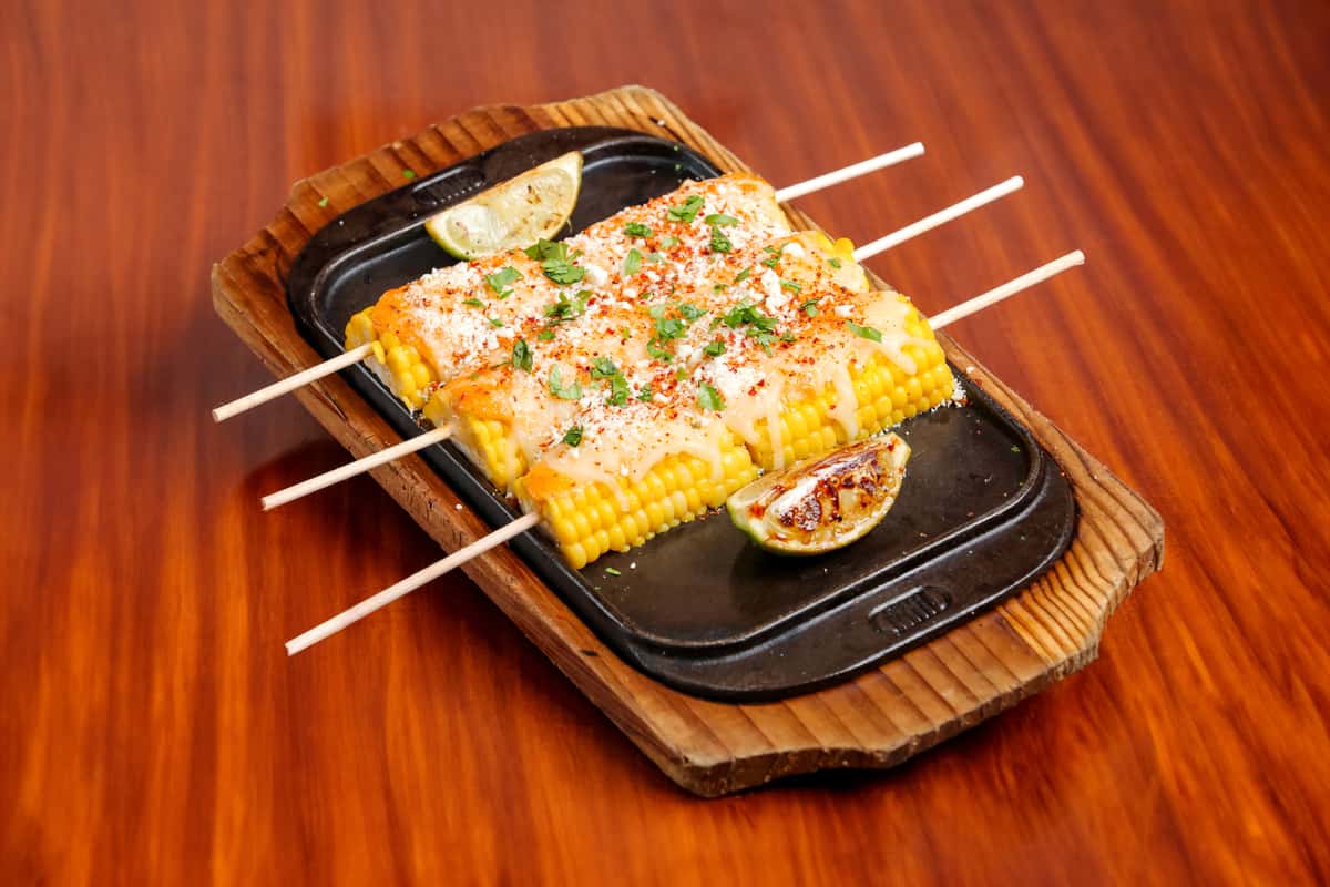 Grilled Mexican street corn on skewers, smothered in melted cheese, cotija, chili powder, and fresh cilantro, served on a sizzling platter with charred lime wedges.
