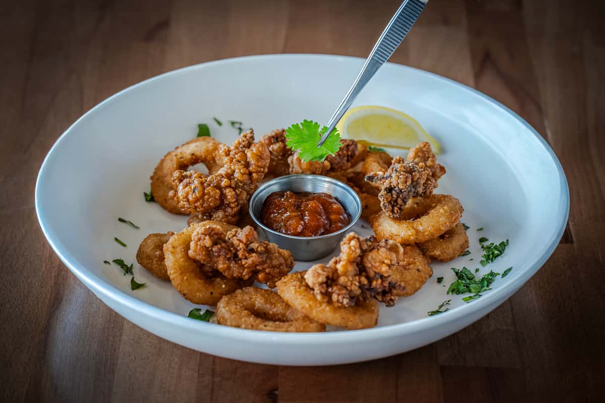 Crispy fried calamari rings and seasoned fried oysters served with a side of zesty marinara sauce, garnished with chopped parsley and a lemon wedge on a white plate.