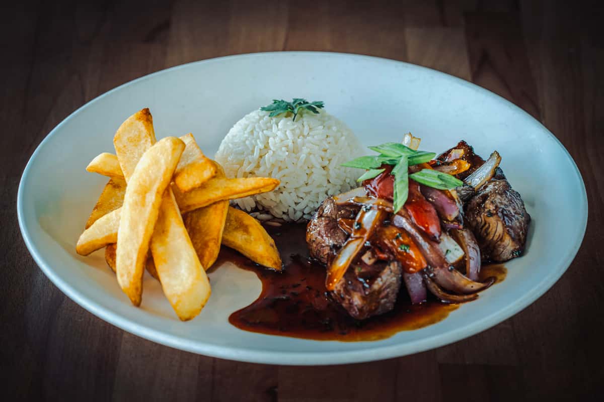 Peruvian-style lomo saltado featuring seared beef strips sautéed with onions and tomatoes in a savory sauce, served with thick-cut fries and a dome of white rice garnished with fresh herbs.
