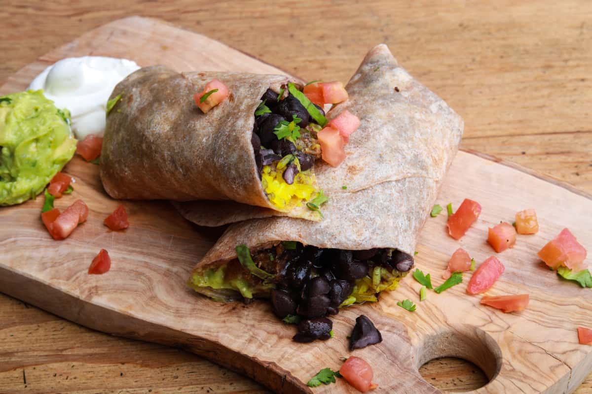 A burrito filled with black beans, yellow rice, and possibly guacamole, served with sour cream and more guacamole on the side. Garnished with chopped tomatoes and fresh cilantro.