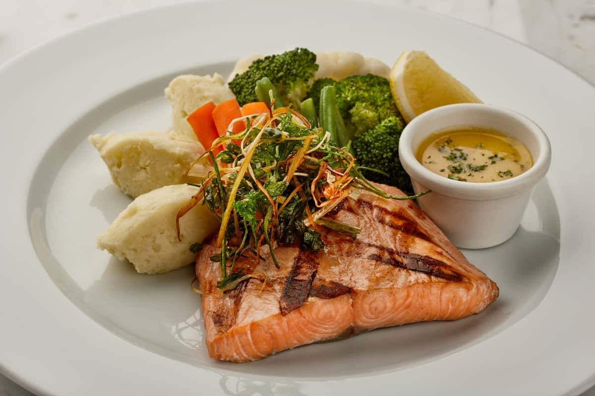 Chargrilled salmon fillet served with mashed potatoes, steamed vegetables, crispy herbs, and a side of creamy lemon butter sauce.