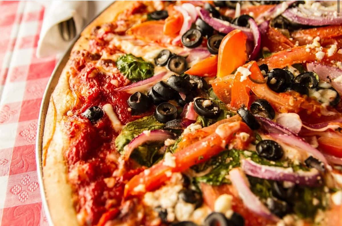 Close-up of a freshly baked vegetarian pizza topped with black olives, red onions, spinach, tomatoes, and crumbled cheese on a golden crust in a cozy Italian restaurant setting.