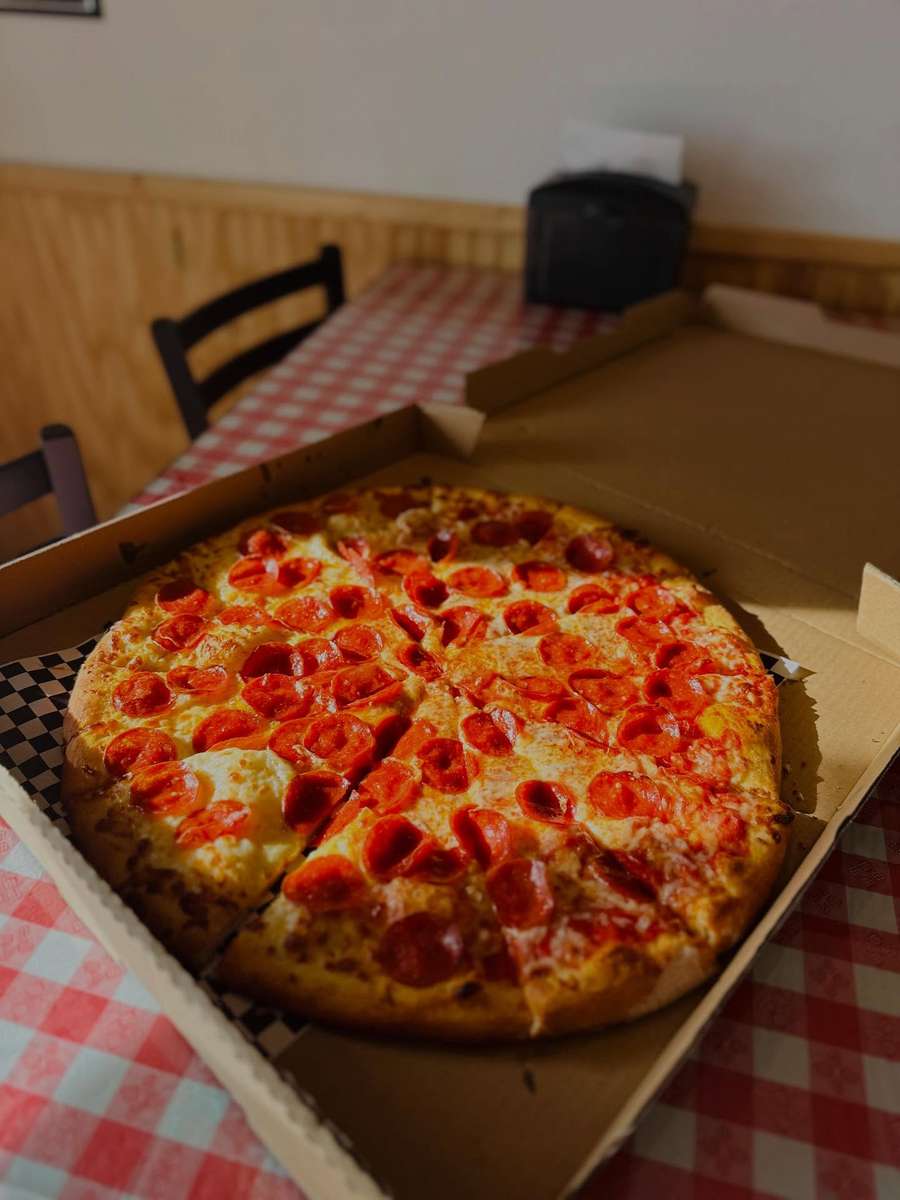 Pepperoni pizza in an open takeout box on a red and white checkered tablecloth, featuring a golden crust, melted mozzarella, and crispy pepperoni slices in a cozy pizzeria setting.