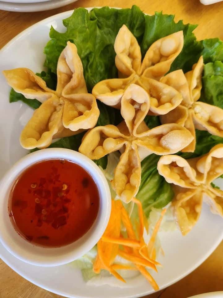 Golden Thai crab rangoon wontons arranged in a flower shape on a bed of fresh lettuce, served with a side of sweet chili dipping sauce and shredded carrots—perfect as a crispy and creamy Thai appetizer.