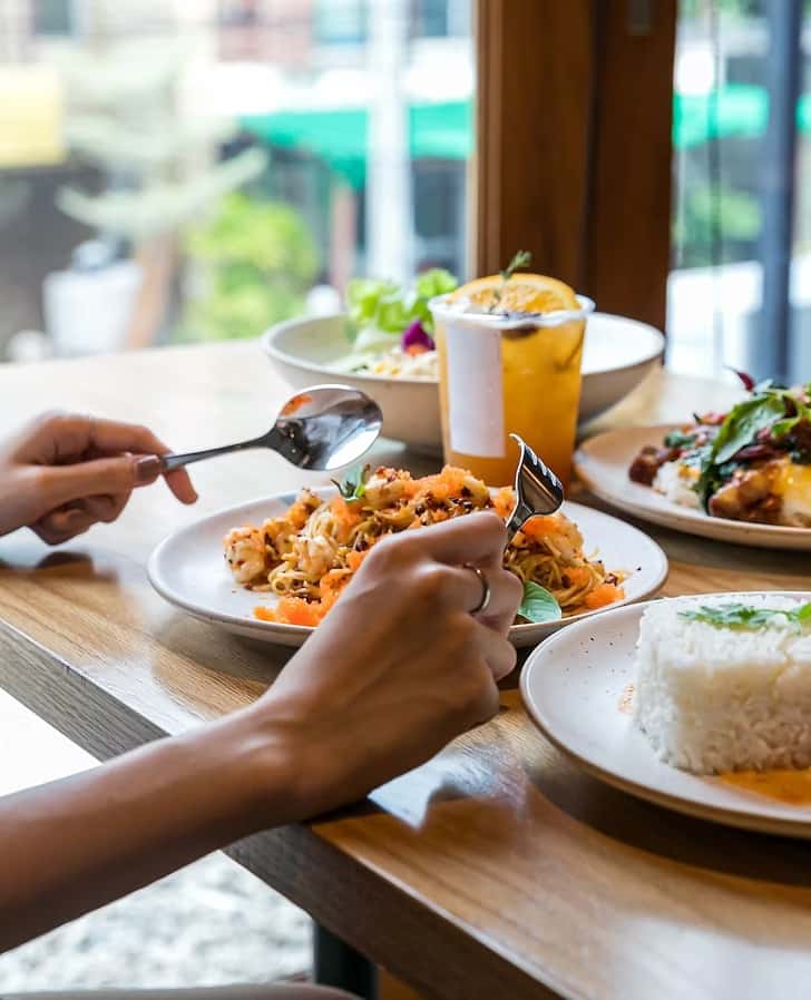 Close-up of a diner enjoying Thai cuisine at a restaurant table, featuring a plate of Pad Thai noodles, jasmine rice, vibrant salads, and a refreshing iced Thai tea with orange slice—set against a bright window with a modern, urban backdrop.