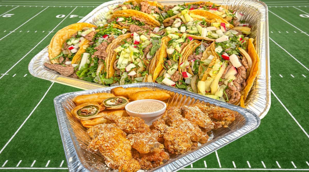 Two aluminum trays of food on a football field background. Top tray: Tacos with beef, cilantro, radish, and cucumber. Bottom tray: Fried wings, jalapeño poppers, and dipping sauce.