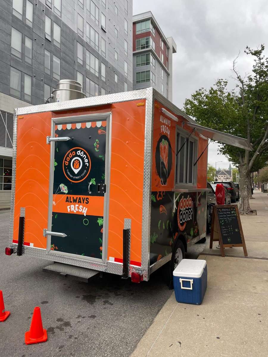 Bright orange Dodo Döner food truck parked on a downtown street with an open service window, outdoor menu board, and modern city buildings in the background.