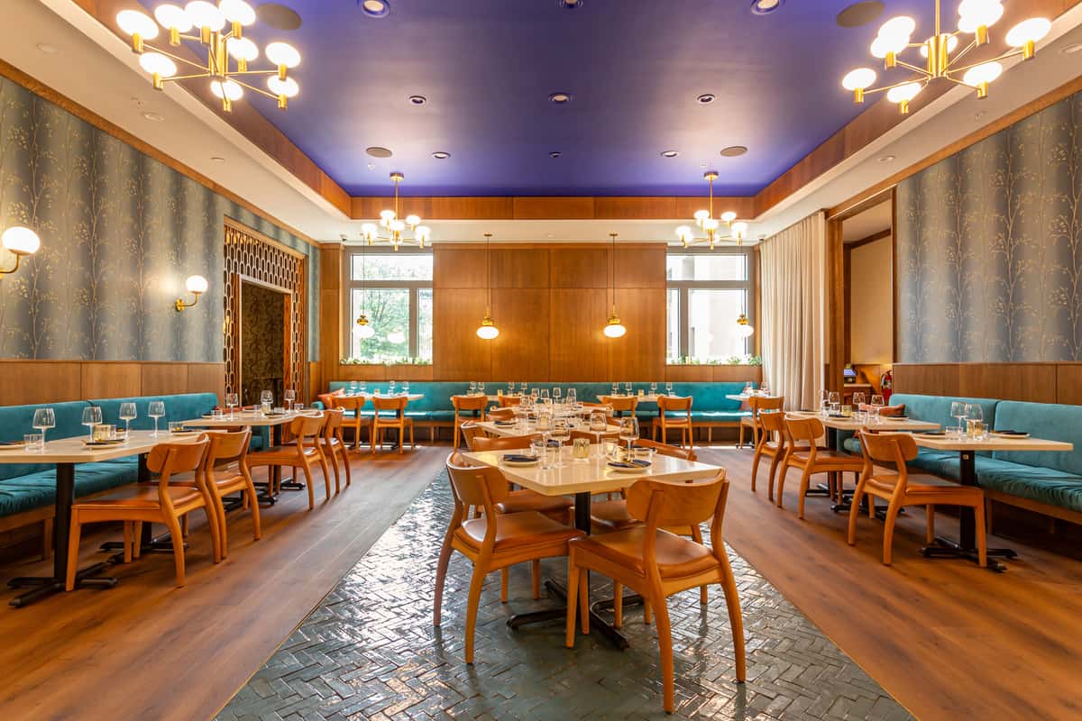Elegant Persian restaurant dining room featuring teal banquettes, light wood chairs, marble tables, and gold-accented lighting under a bold royal blue ceiling, creating a warm and contemporary Middle Eastern ambiance.