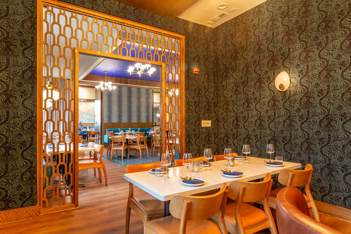 "Chic Persian restaurant interior featuring a semi-private dining area with ornate wooden lattice partition, elegant wallpaper, and a white table set for six with modern wooden chairs and blue-accented place settings.