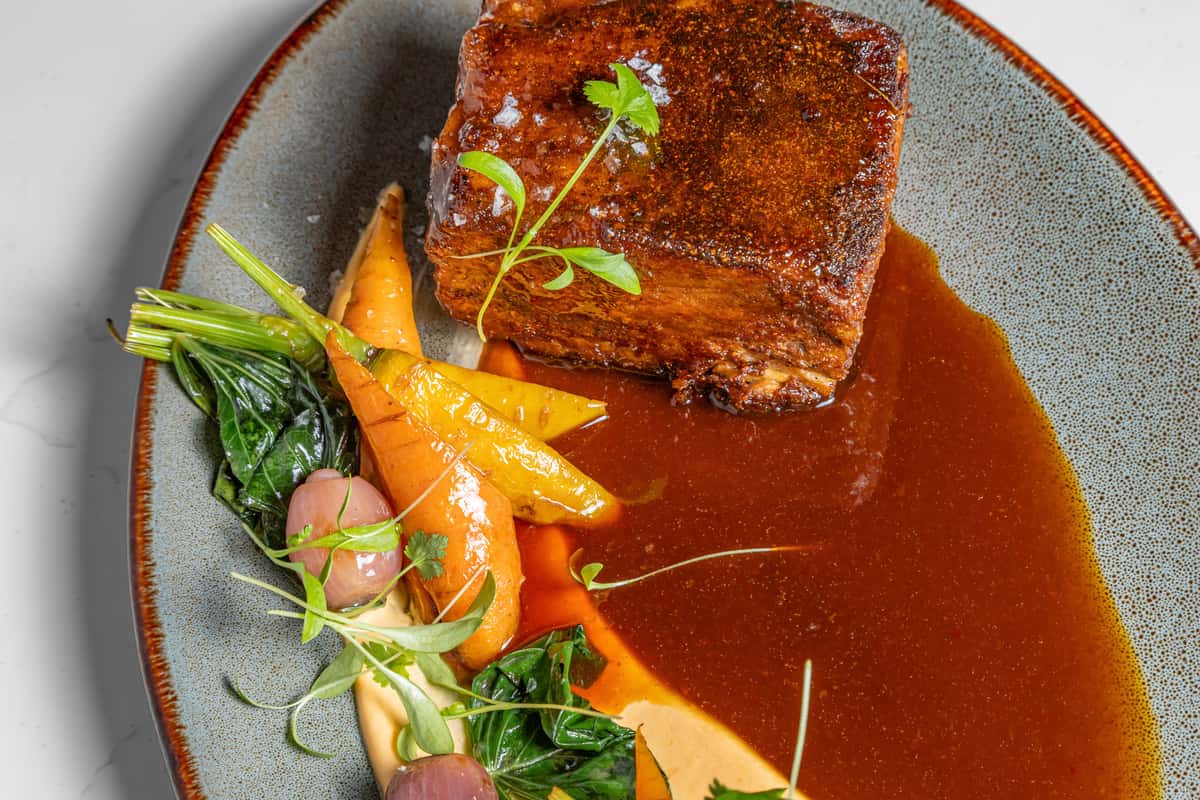 Tender braised short rib glazed with rich demi-glace, served with heirloom carrots, sautéed greens, and pickled onions over creamy puree on a speckled ceramic plate.
