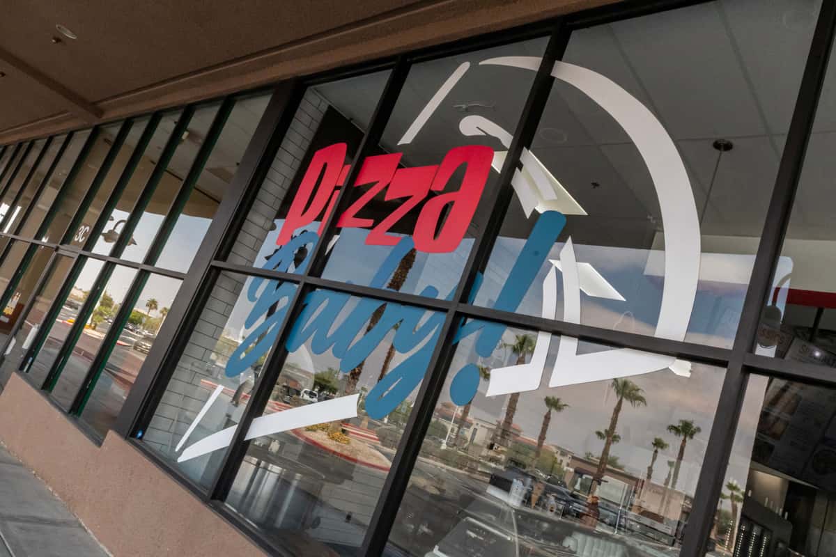 Exterior window of Pizza Baby! restaurant with bold red, blue, and white branding on large glass panels, reflecting palm trees and nearby shops for a vibrant, modern pizzeria look in a sunny shopping plaza setting.