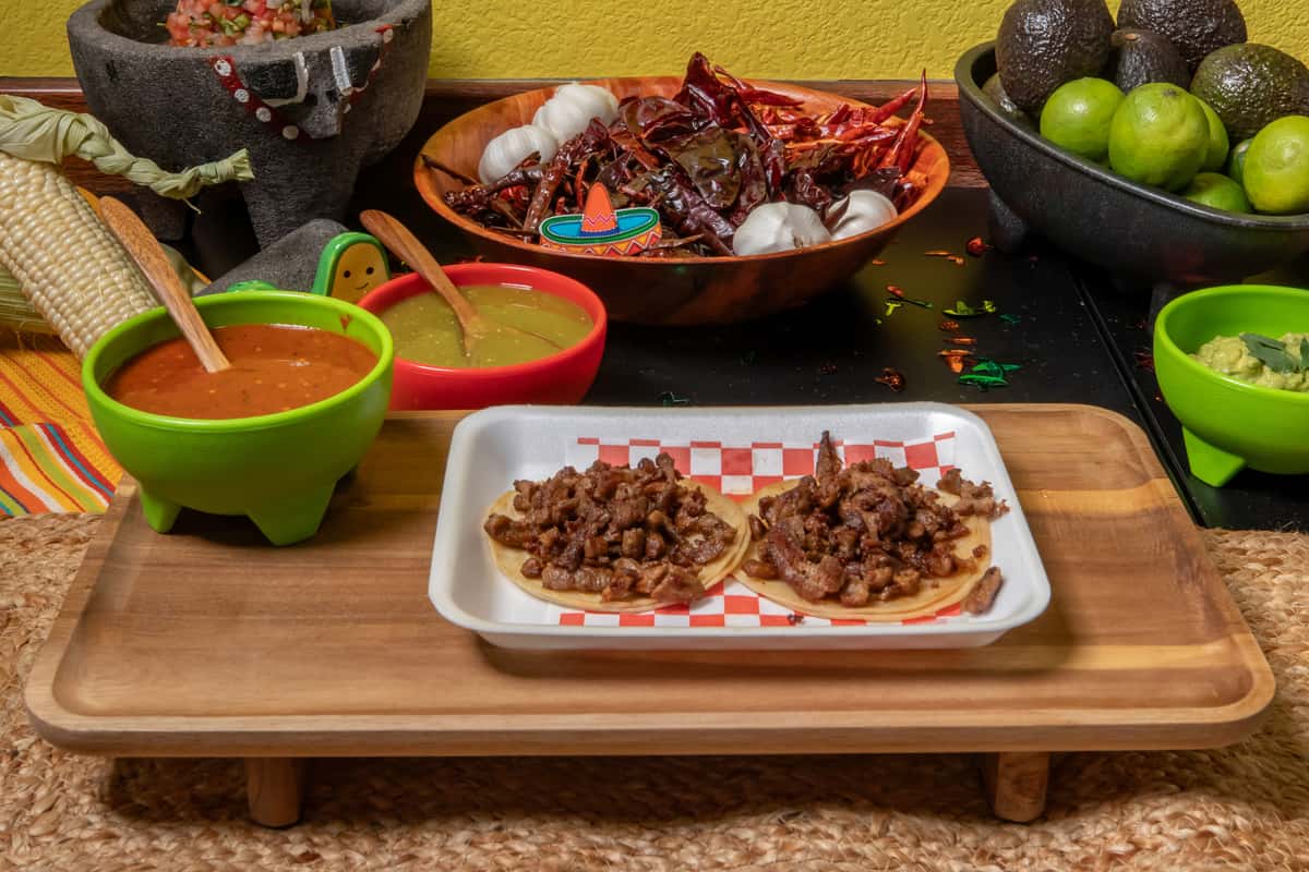 Two carne asada street tacos served on corn tortillas, piled with grilled and seasoned beef. Presented on a red checkered tray alongside traditional Mexican condiments—red salsa, green salsa, guacamole—and surrounded by dried chiles, garlic, avocados, and limes for an authentic taqueria ambiance.