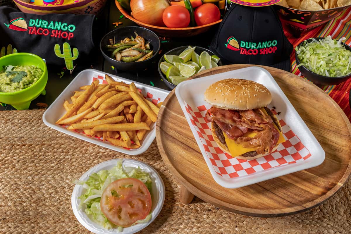 Juicy bacon cheeseburger with crispy bacon strips and melted cheddar on a sesame seed bun, served with seasoned French fries and a side of lettuce and tomato. Displayed in a festive taqueria setting with guacamole, sautéed peppers, and Durango Taco Shop branding in the background.