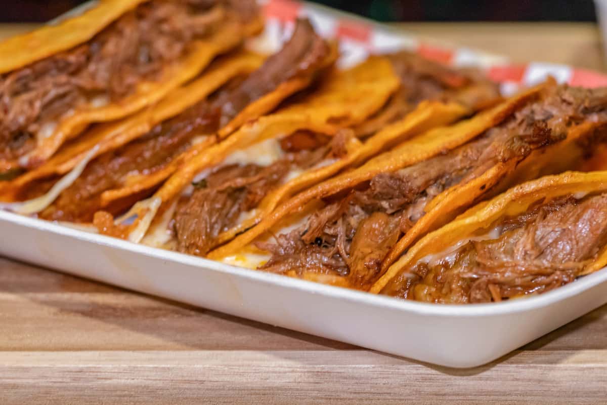 Golden, crispy birria tacos stuffed with tender, slow-cooked shredded beef and melted cheese, lined up in a tray. The tacos have a rich, reddish hue from the flavorful consommé, offering a bold and savory street food presentation.