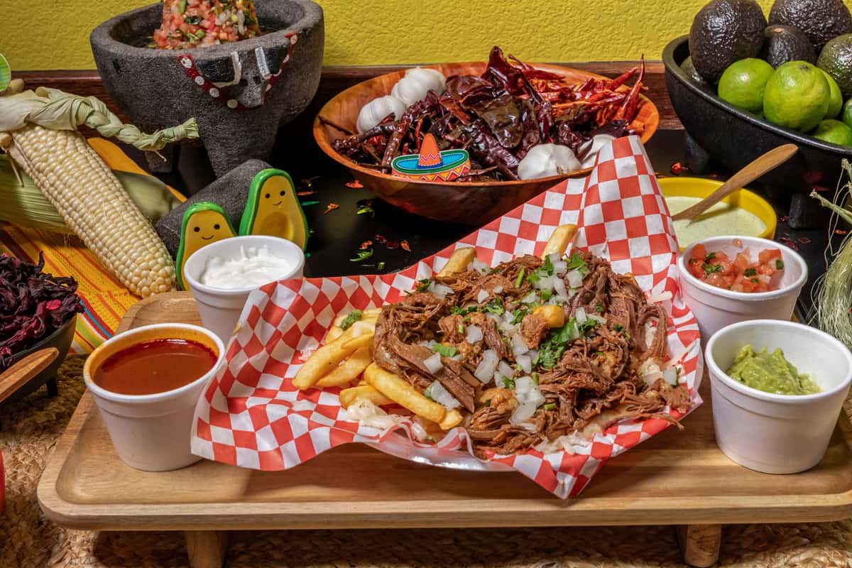 Birria-loaded fries topped with tender shredded beef, chopped onions, and cilantro, served on a red checkered tray with sides of guacamole, sour cream, pico de gallo, and a cup of rich consommé. Surrounded by traditional ingredients like dried chiles, garlic, corn, and limes, evoking authentic Mexican street food flair.