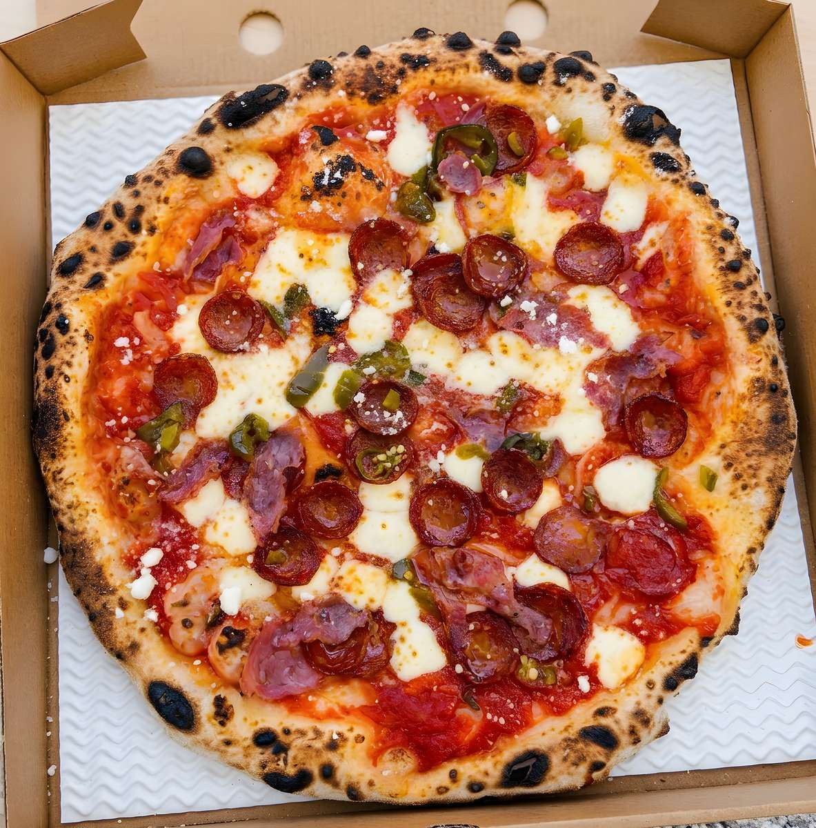 Neapolitan pizza with pepperoni, salami, green peppers, mozzarella, and a charred crust in a box.