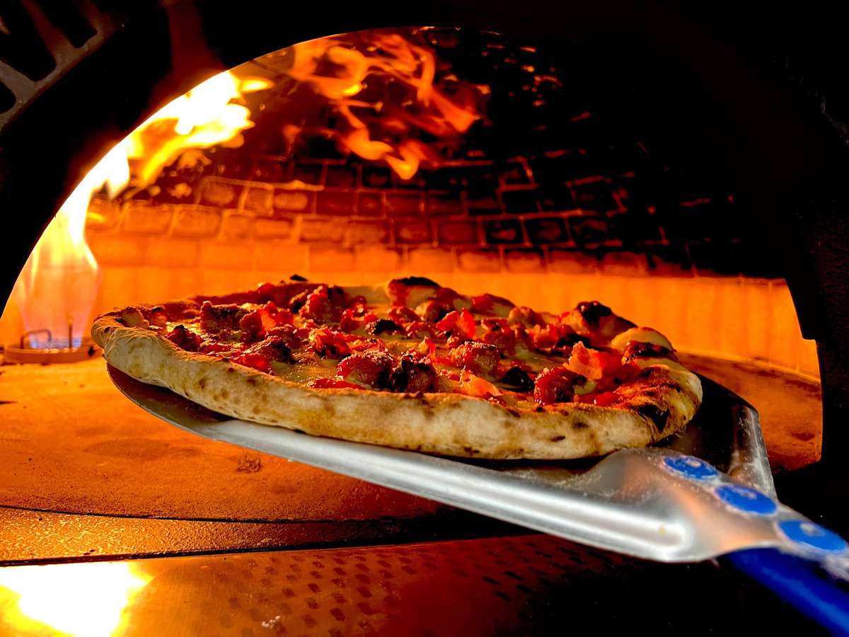 Pizza baking in a brick oven with a blue gas flame visible. A metal pizza peel rests in the foreground.