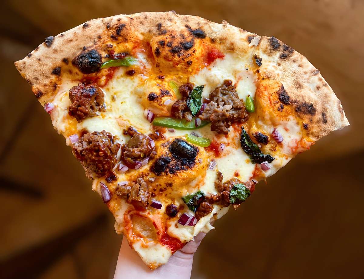 Slice of pizza with melted cheese, tomato sauce, green bell peppers, red onions, crumbled meat, and dark herbs held in a hand.