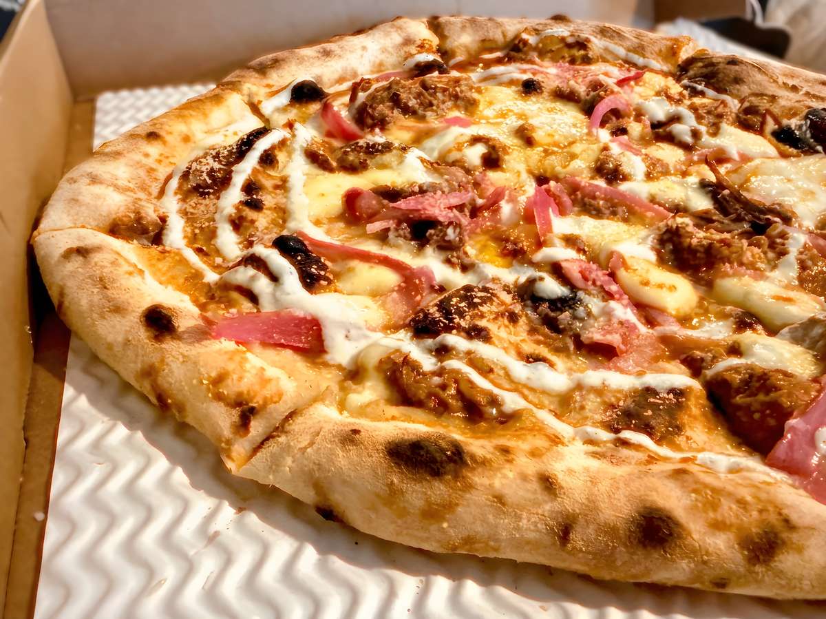 Pizza with melted cheese, pulled pork, pickled red onions, and a white sauce drizzle in a delivery box.