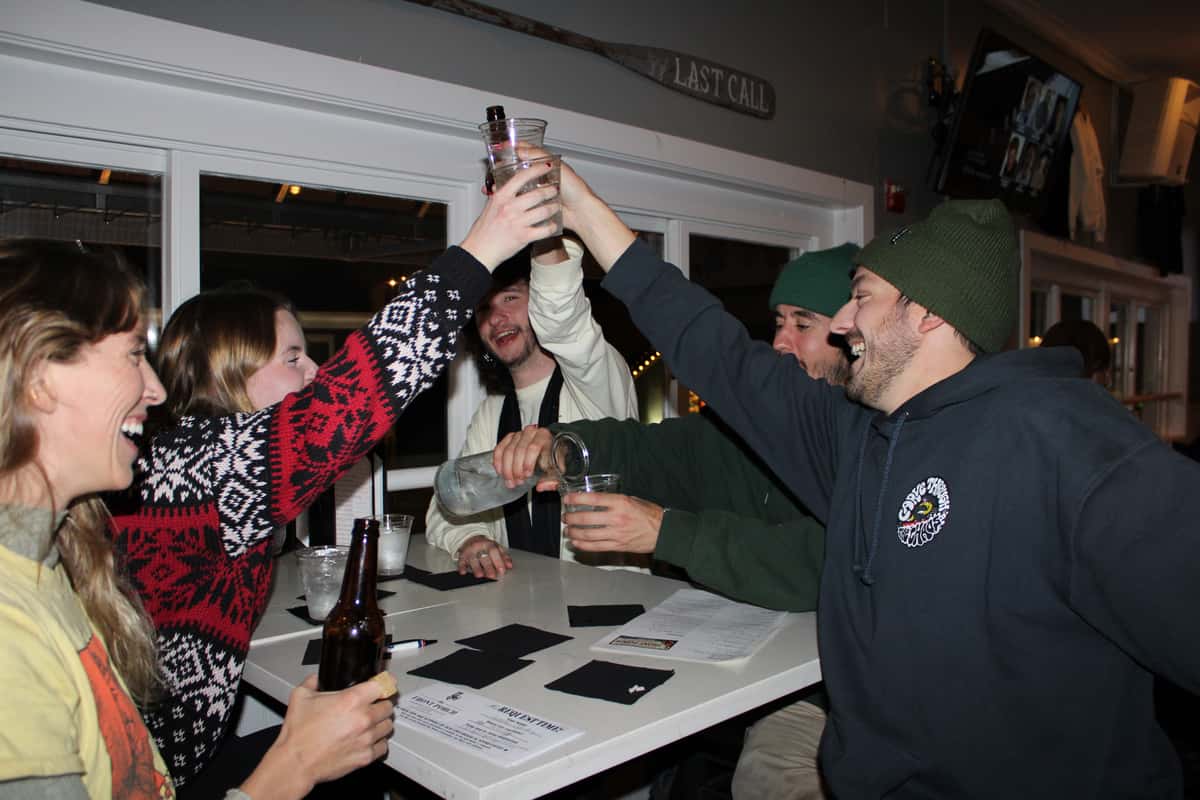 Group of friends laughing and raising drinks for a toast around a bar table with black napkins and printed menus.