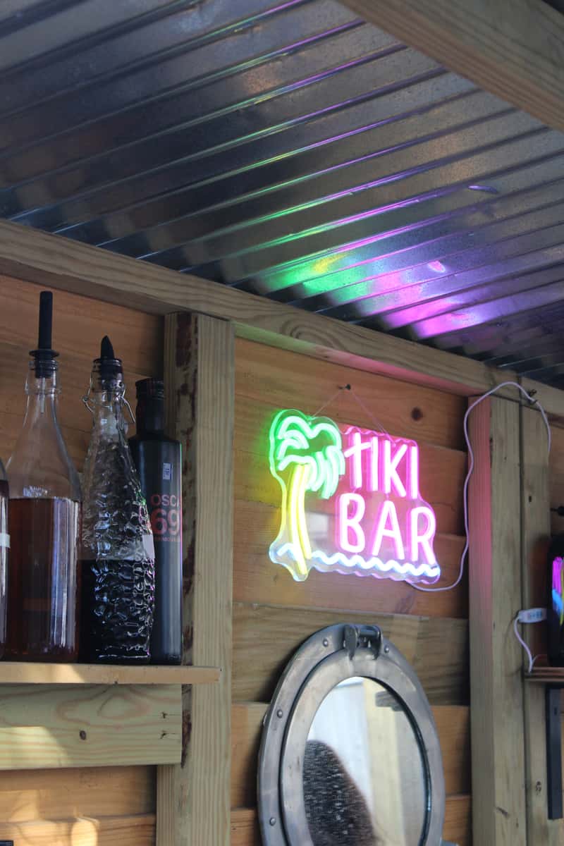 Neon "Tiki Bar" sign glows on a wooden wall beside bottles and a round porthole-style mirror under a metal roof.Neon "Tiki Bar" sign glows on a wooden wall beside bottles and a round porthole-style mirror under a metal roof.